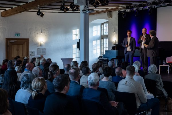 3 Herren stehen auf einer Bühne vor Publikum im Gespräch